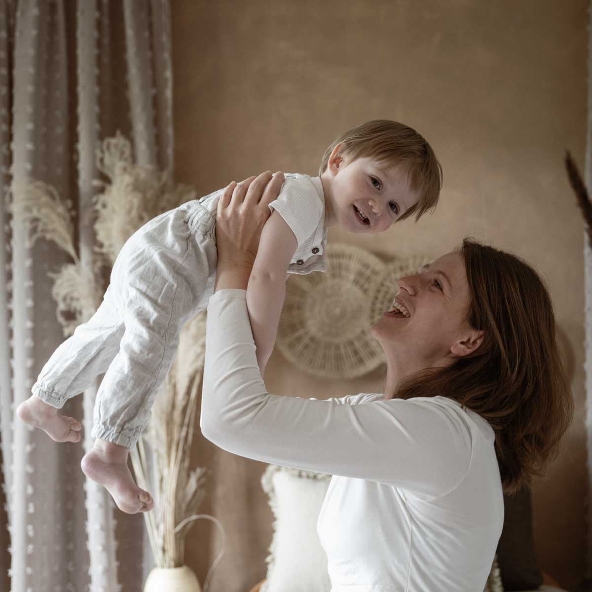 Mutter hebt ihr lachendes Kleinkind spielerisch in die Höhe in einem warm dekorierten Raum mit Naturtönen.